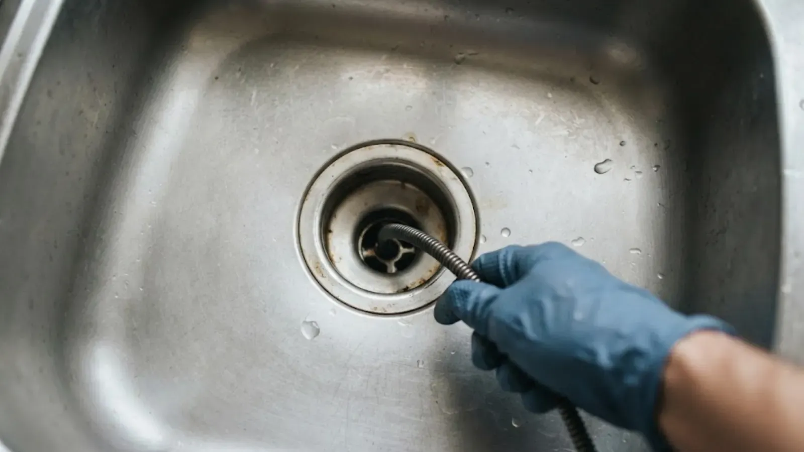 Gloved hand feeding a drain cable into a stainless steel sink drain